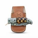 Engraved Bolster Damascus Folding Knife with patterned curved blade, brown and black checkered handle, floral metal fittings and brown stitched sheath.