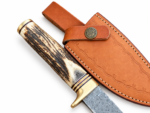 Patterned gut hook knife with a dark antler-look handle, cream ends, and gold-tone fittings resting on a tan stitched sheath.