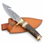 Patterned gut hook knife with a dark antler-look handle, cream ends, and gold-tone fittings resting on a tan stitched sheath.