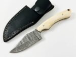 Ivory Handle Curved Skinner Knife with silver-gray Damascus-style patterned blade, cream-colored contoured handle and black stitched leather-style sheath.