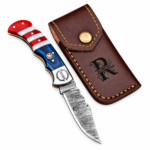 Open red white and blue folding knife with a patterned blade, star handle detail and monogrammed silver-tone bolster resting on a brown stitched pouch.