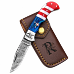 Open red white and blue folding knife with a patterned blade, star handle detail and monogrammed silver-tone bolster resting on a brown stitched pouch.