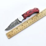 Red grain handle folding knife shown open beside a ruler, with a gray patterned blade, silver-tone bolster, and red-and-black contoured grip.