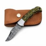 Basket weave handle folding knife with a gray patterned blade, silver-tone bolster, and black-and-gold handle resting across a brown snap-fastened pouch.