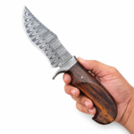 Brown handle Bowie knife held in a hand, showing a broad gray patterned blade, matching curved guard, hooked grain-look grip, and silver-tone pin.