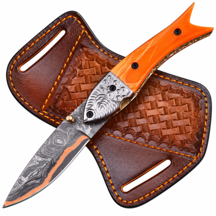 Orange fishtail folding knife with a patterned blade, copper-tone stripe, and ornate silver-tone bolster resting on a brown basketweave-style sheath.