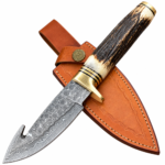 Patterned gut hook knife with a dark antler-look handle, cream ends, and gold-tone fittings resting on a tan stitched sheath.