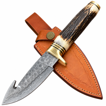 Patterned gut hook knife with a dark antler-look handle, cream ends, and gold-tone fittings resting on a tan stitched sheath.