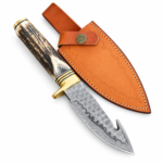 Patterned gut hook knife with a dark antler-look handle, cream ends, and gold-tone fittings resting on a tan stitched sheath.