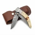 Damascus Folding Pocket Knife shown open with a patterned silver-gray blade, gold-colored thumb stud, cream bone-style handle and decorative metal inlay.