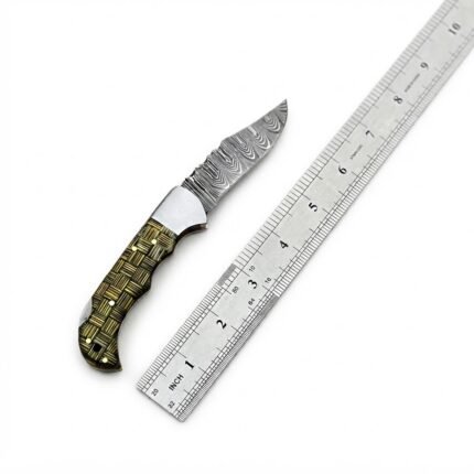 Basket weave handle folding knife with a gray patterned blade, silver-tone bolster, and black-and-gold handle resting across a brown snap-fastened pouch.