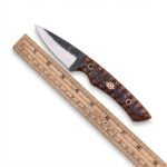 Patterned handle skinner knife with dark and silver pointed blade, brown-and-black decorative grip, mosaic-style pin and black sheath with tan stitching.